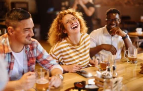 Imagem de três amigos sentados em uma mesa de bar de madeira, rindo e saboreando canecas de cerveja. A mulher no centro está usando uma camisa listrada e está rindo alegremente. Pratos de comida podem ser vistos em primeiro plano.