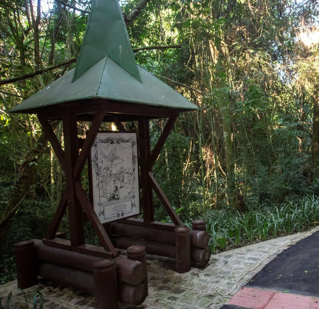 Imagem da entrada do Caminho do Conto, no Bosque Alemão, em Curitiba, mostra uma grande placa informativa de madeira com um telhado verde triangular em um caminho pavimentado cercado por uma densa floresta e vegetação. A placa exibe uma ilustração em preto e branco.