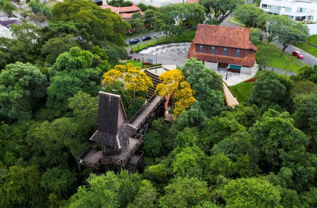 Imagem da vista aérea da Torre dos Filósofos, no Bosque Alemão, mostra uma casa na árvore de madeira cercada por densas árvores verdes, com um prédio de telhado vermelho e um estacionamento ao fundo.