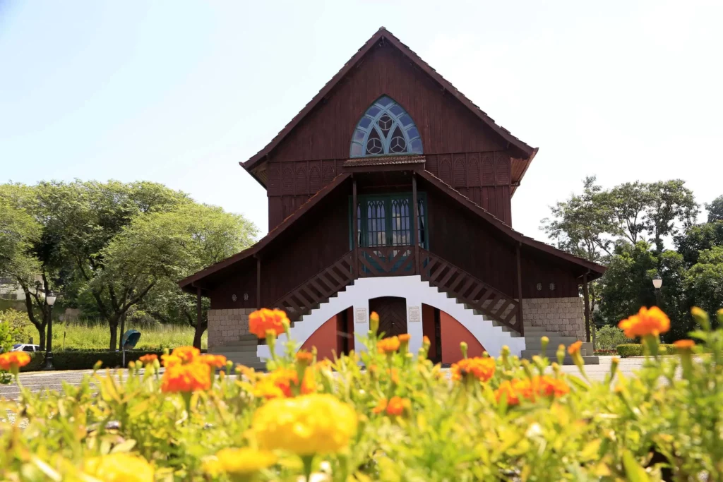 Imagem do Oratório Bach, no Bosque Alemão, mostra uma casa de madeira marrom com janelas em arco e degraus brancos que fica atrás de um jardim de flores amarelas e laranjas vibrantes sob um céu claro. Árvores e vegetação cercam o edifício.