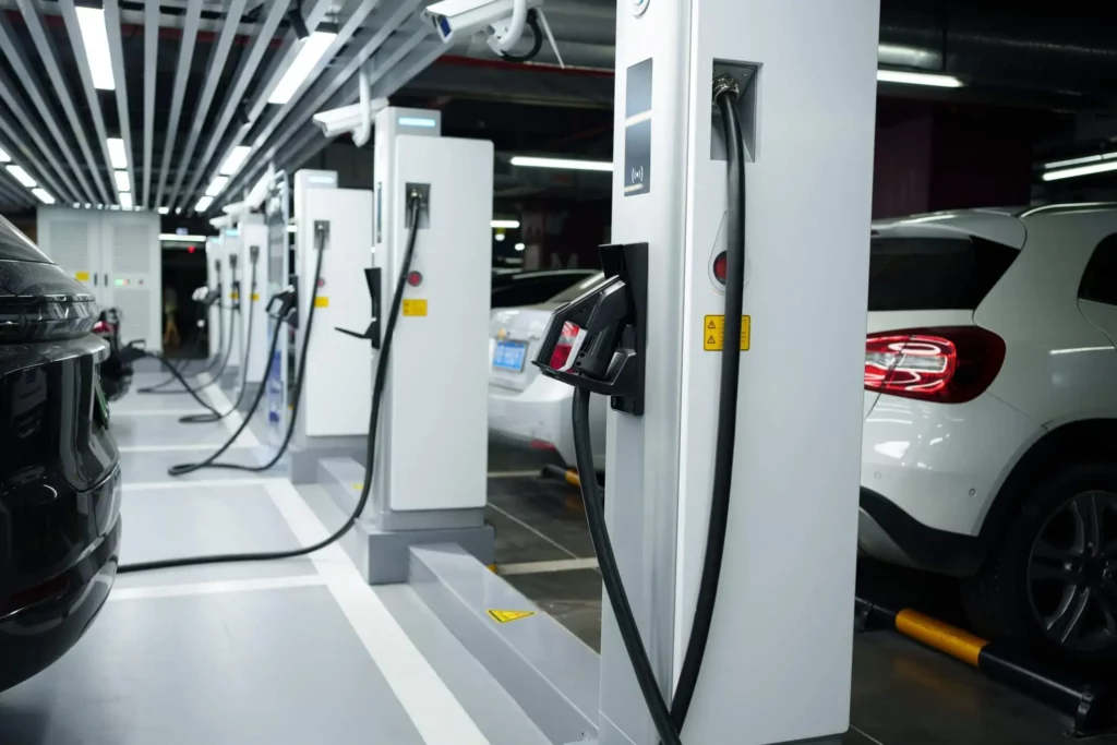 Imagem de uma fileira de estações de carregamento de veículos elétricos em uma área de estacionamento coberta, com vários carros estacionados e conectados aos carregadores. A área é bem iluminada com luzes no teto.