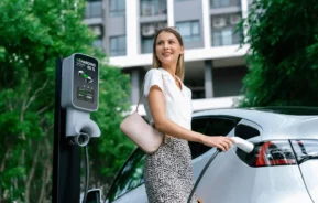 Imagem de uma mulher ao lado de um carro elétrico branco, segurando um plugue de carregamento em uma estação de carregamento ao ar livre. Ela está sorrindo e parece relaxada, com árvores e um edifício moderno ao fundo.