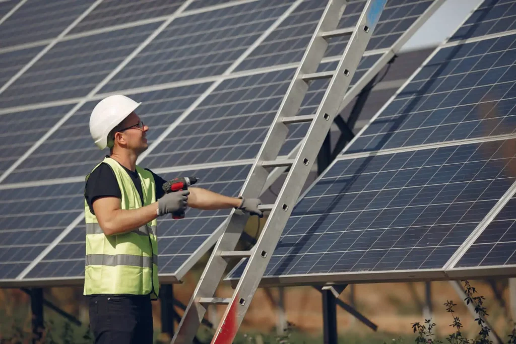 Imagem de um trabalhador com colete de segurança e capacete ao lado de uma escada apoiada em grandes painéis solares, segurando uma furadeira elétrica e parecendo inspecionar ou realizar a manutenção do painel solar para condomínios ao ar livre.