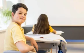 Imagem de um garoto de camisa amarela sentado em uma carteira escolar, sorrindo e olhando para a câmera. Outro aluno com cabelo comprido olha para frente em uma carteira próxima. Materiais escolares e papéis estão sobre as carteiras.