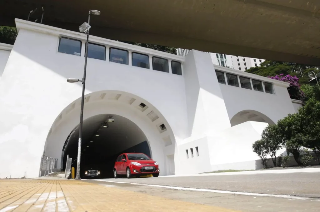 Imagem de um carro vermelho saindo do Túnel 9 de Julho, em São Paulo, na Avenida Nove de Julho. Outro carro segue atrás. O túnel é cercado por árvores e edifícios modernos, com uma faixa de pavimentação tátil amarela em primeiro plano.
