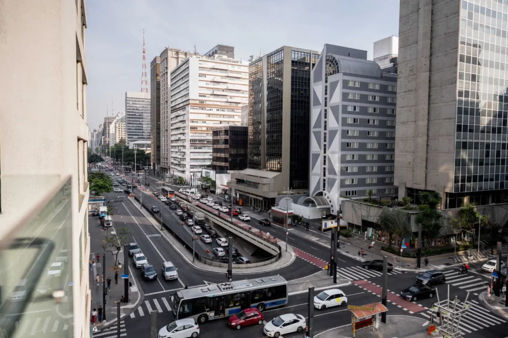 Imagem da vista aérea da Avenida Paulista, que fica perto da Avenida Nove de Julho, em São Paulo, edifícios altos e modernos. Algumas áreas verdes revestem as calçadas e o tráfego se move em várias direções. O céu está ligeiramente nublado.