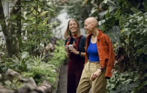 Imagem de duas mulheres caminhando e rindo em uma estufa; uma segura uma câmera, enquanto a outra usa uma jaqueta vermelha e um top azul. Elas estão cercadas por plantas verdes e parecem estar aproveitando o momento juntas