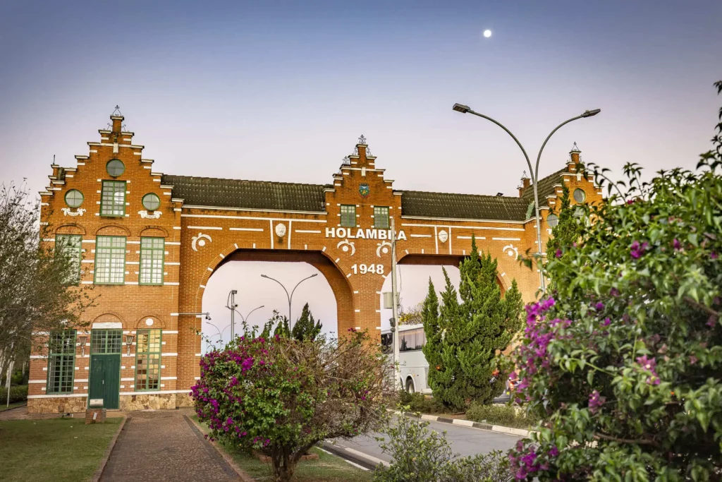 Imagem de um grande arco de tijolos ornamentado em uma estrada na entrada de Holambra cercado por árvores e arbustos floridos sob um céu claro com a lua visível acima.