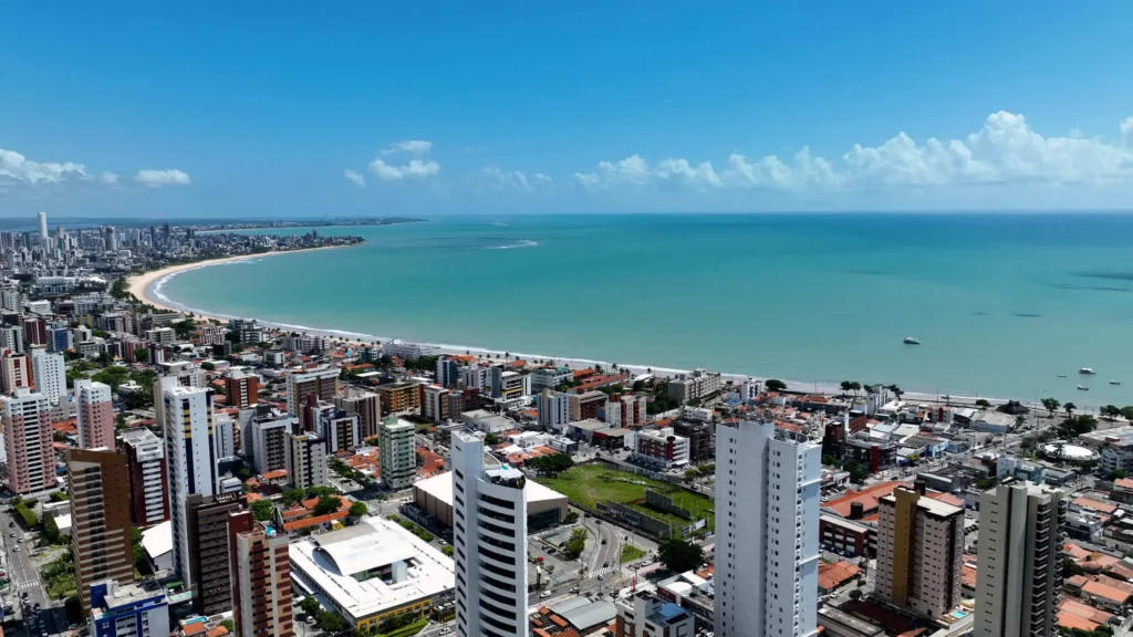 Imagem da vista aérea de João Pessoa mostra prédios altos, casas com telhados vermelhos e uma longa praia de areia curvada ao longo do mar azul-turquesa sob um céu azul claro.