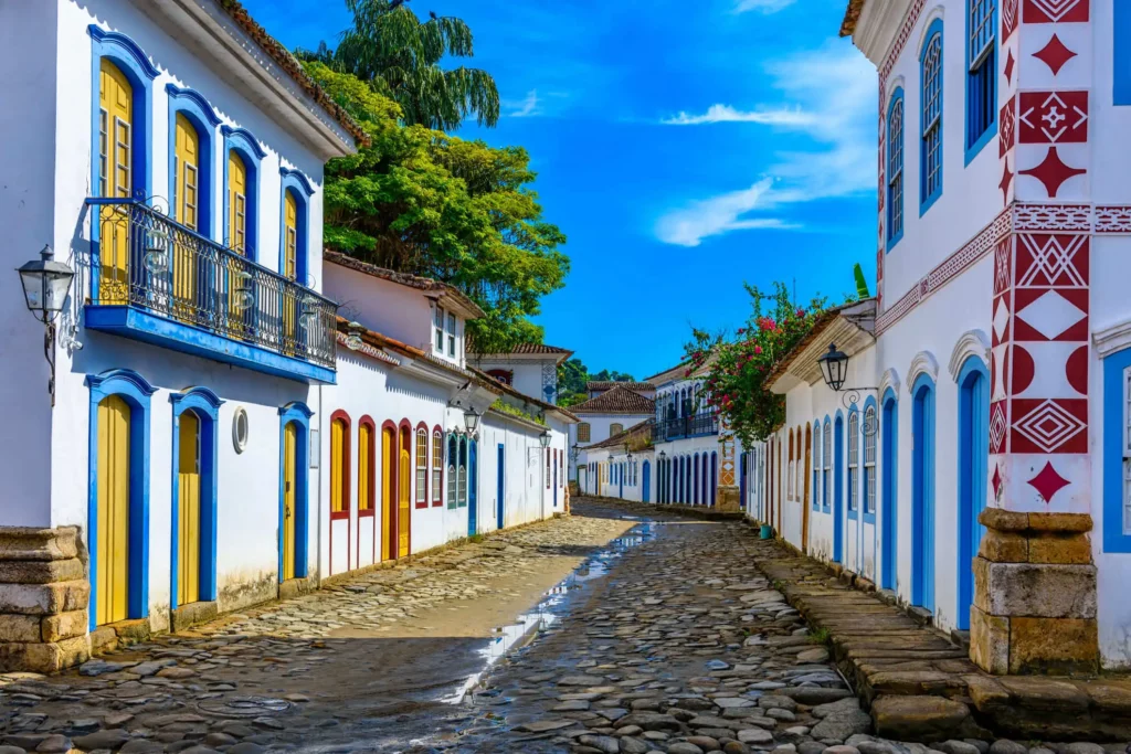 Imagem de prédios coloridos em estilo colonial, com varandas e portas de madeira, alinham-se em uma rua de paralelepípedos sob um céu azul brilhante em Paraty, no Rio de Janeiro. Árvores verdes exuberantes são visíveis ao fundo