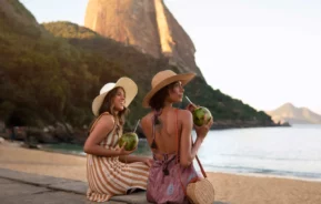 Imagem de duas mulheres com vestidos e chapéus de verão sentadas em uma praia, sorrindo e segurando cocos. Há montanhas ao fundo e água calma ao lado delas, sugerindo um ambiente relaxante e tropical.