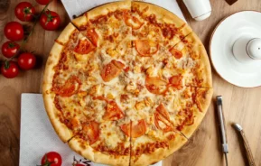 Imagem de uma pizza inteira de queijo coberta com fatias de tomate e pedaços de atum, cortada em oito pedaços, em uma mesa de madeira ao lado de tomates, talheres e um prato branco.