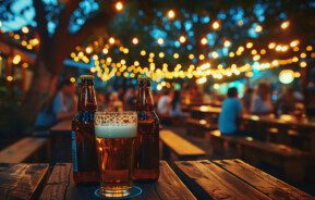 Imagem de um copo e duas garrafas de cerveja sobre uma mesa de madeira em um bar ao ar livre ao anoitecer, com luzes de corda brilhando no alto e pessoas sentadas em mesas de piquenique ao fundo.