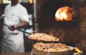 Imagem de um chef em um uniforme branco deslizando uma pizza recém-assada de um forno a lenha para uma pilha de pratos, com as chamas do forno brilhando ao fundo, para ilustrar matéria sobre as melhores pizzarias de SP.