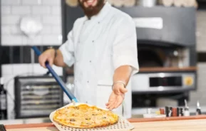 Imagem de um chef em um uniforme branco sorrindo enquanto segura uma pizza, apresentando-a em uma cozinha com um forno ao fundo.