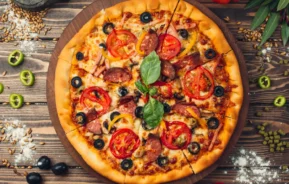 Imagem de uma pizza recém-assada coberta com tomates, linguiça, azeitonas e manjericão em uma tábua de madeira. Ao redor dela, há legumes fatiados, azeitonas, farinha e ervas, todos dispostos em uma mesa de madeira rústica.