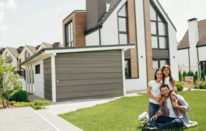 Imagem de uma família sorridente de três pessoas sentada na grama em frente a uma casa moderna com grandes janelas e uma garagem. Os pais estão sentados e a filha está atrás deles, todos parecendo felizes e relaxados.