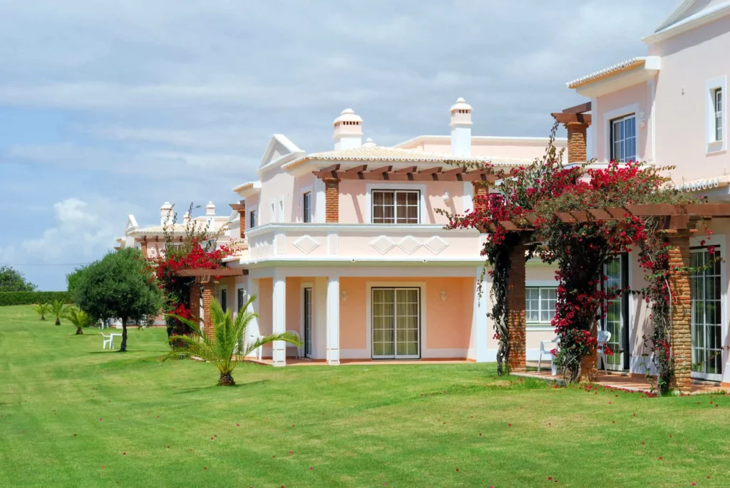 Imagem de uma casa clássica cor de pêssego com acabamento e varandas brancas, cercadas por grama verde e videiras floridas, sob um céu nublado.