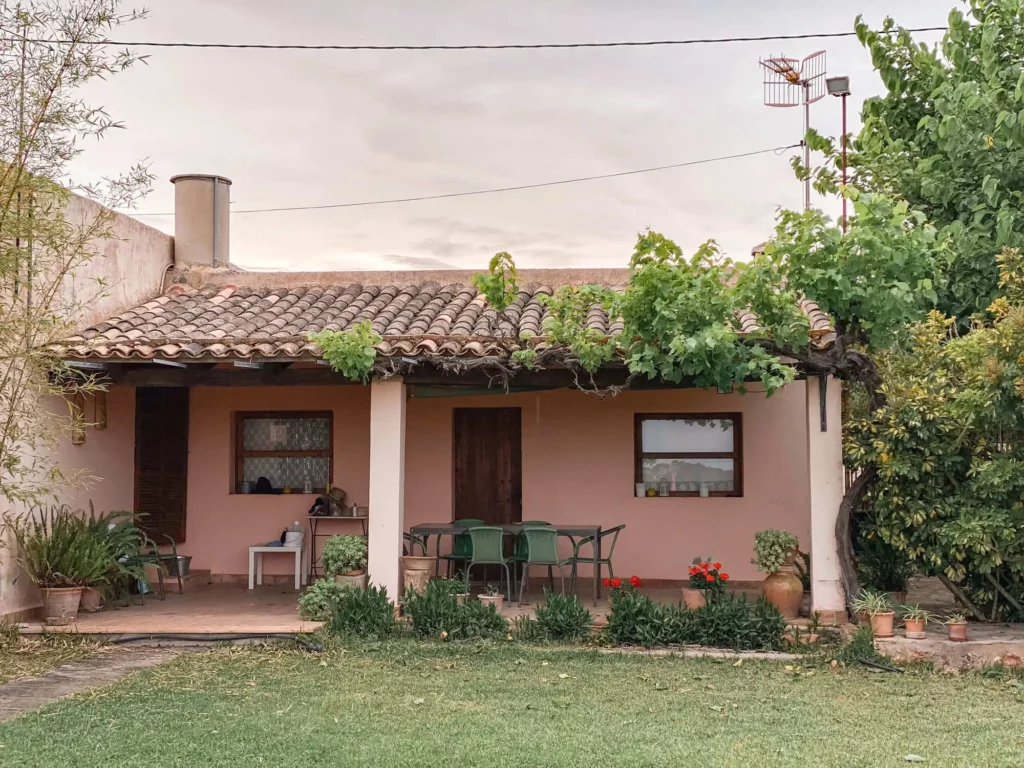  Imagem de uma casa de alvenaria com uma varanda coberta, com cadeiras verdes e uma mesa. Vasos de plantas e flores estão alinhados na frente, e uma árvore cresce em uma coluna. O gramado é verde e bem cuidado.