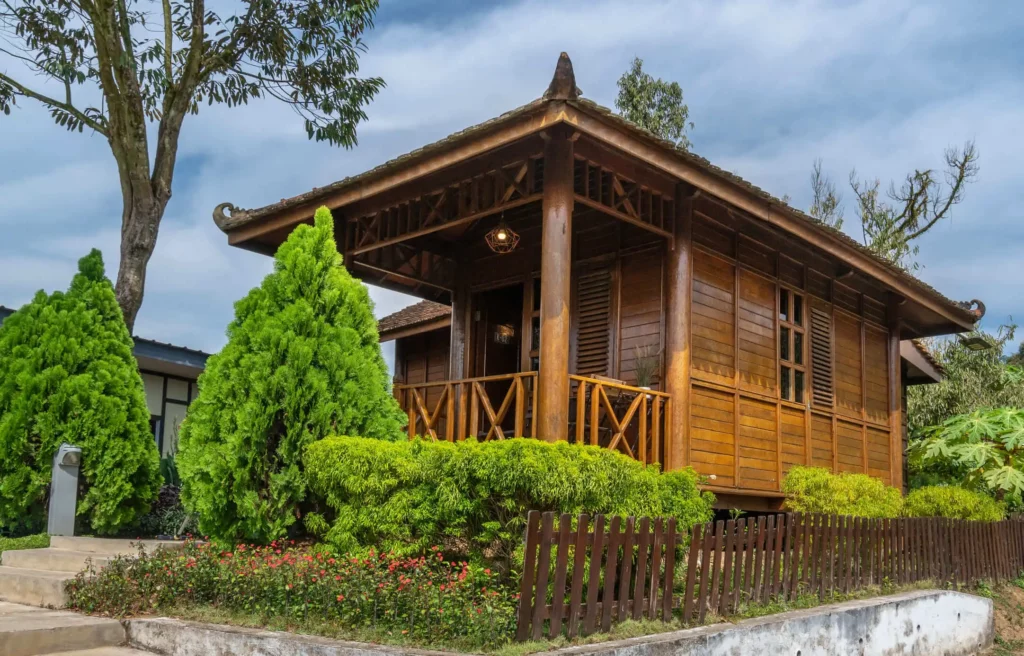 Imagem de uma casa de madeira com varanda e corrimão na frente, cercada por arbustos verdes, árvores e uma cerca de madeira, sob um céu parcialmente nublado.