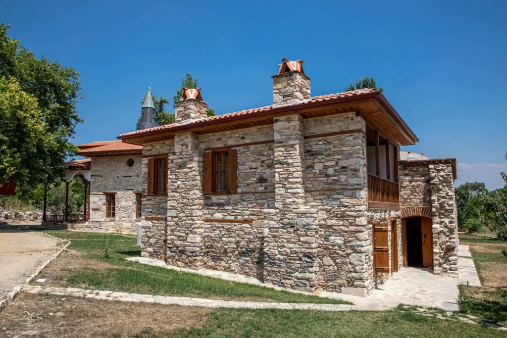 Imagem de uma casa de pedra com janelas de madeira e telhado de telhas vermelhas em um terreno gramado, sob um céu azul claro. Duas chaminés são visíveis no telhado, e um caminho pavimentado leva à entrada. As árvores cercam o imóvel.