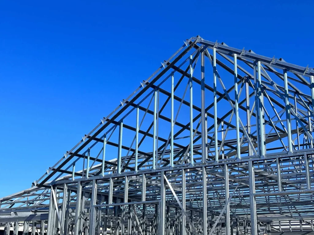 Imagem de estrutura de aço de uma casa de steel frame em construção contra um céu azul claro, mostrando a forma triangular do telhado e várias vigas e suportes.
