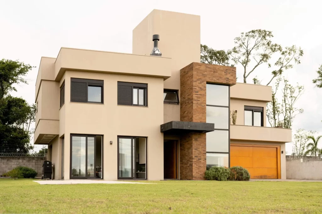 Imagem de uma casa duplex moderna com paredes bege, janelas grandes, uma seção de tijolos, persianas pretas e uma porta de garagem laranja brilhante, situada em um gramado verde com algumas árvores ao fundo.