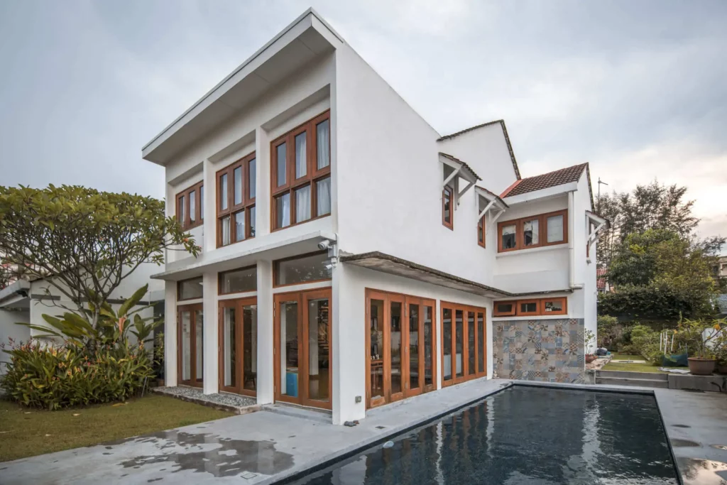 magem de uma casa em L moderna, branca e de dois andares com grandes janelas e portas com estrutura de madeira, cercada por vegetação e uma piscina retangular no quintal sob um céu nublado.