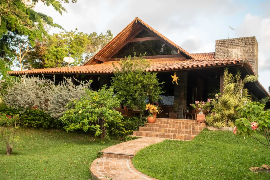 Imagem de uma casa rústica com chaminé de pedra em meio a um gramado com um caminho de tijolos que leva a uma varanda coberta cercada por várias plantas e árvores.