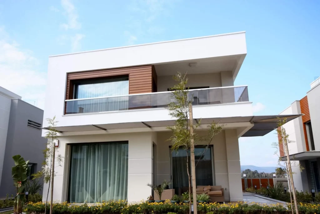 Imagem de um sobrado de dois andares com janelas grandes, uma varanda com guarda-corpo de vidro e um pequeno jardim com flores e árvores jovens no jardim da frente sob um céu claro.