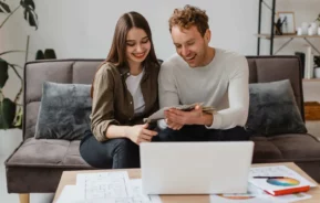 Imagem de um casal sorridente sentado em um sofá, olhando para papéis e um tablet em suas mãos, com um laptop e plantas arquitetônicas espalhadas na mesa de centro à sua frente.