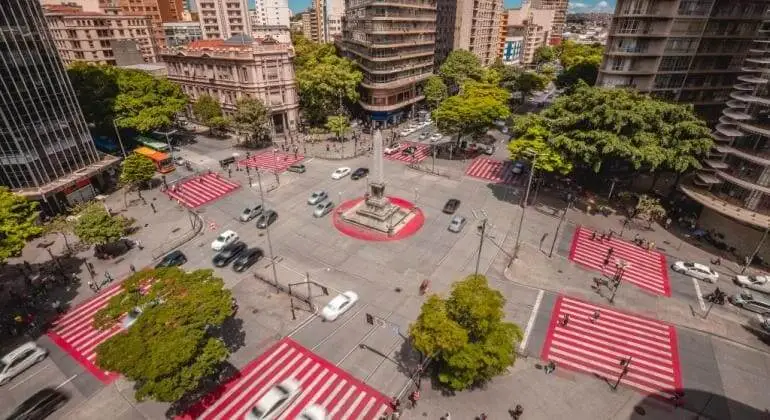 Imagem da vista aérea da Praça 7 de Setembro mostra cruzamentos ao redor do monumento central. A praça está cercada por prédios altos, árvores e faixas de pedestres marcadas com listras vermelhas e brancas. Carros e pedestres circulam pelas ruas urbanas