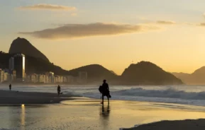 Imagem de um surfista caminhando na praia de Copacabana, no Rio de Janeiro, durante o pôr do sol. Montanhas e prédios urbanos estão ao fundo, além de ondas calmas se aproximando da costa sob um céu amarelo-alaranjado.