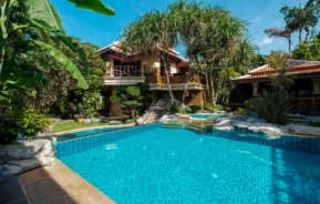 Imagem de uma piscina azul clara cercada por plantas e árvores tropicais, com uma casa de dois andares com varandas e telhados de telha ao fundo, sob um céu azul brilhante.