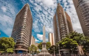 Imagem do Obelisco na Praça Sete de Setembro, em Belo Horizonte, cercado por prédios altos e árvores verdes sob um céu azul brilhante com nuvens brancas espalhadas. As pessoas caminham pelas ruas movimentadas.