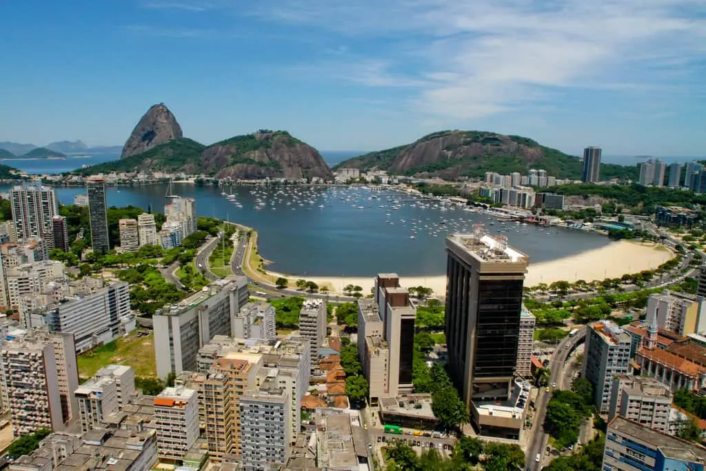 Imagem da vista aérea da Praia de Botafogo, no Rio de Janeiro, mostra prédios altos, uma praia de areia curva e a Baía de Guanabara sob um céu azul.