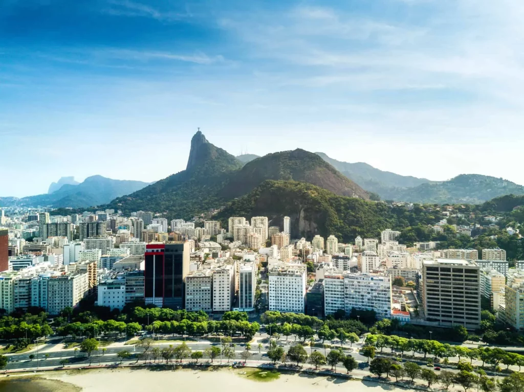 Vista aérea do Rio de Janeiro, Brasil, mostrando prédios altos, colinas verdejantes e o icônico Pão de Açúcar ao fundo, sob um céu azul claro.