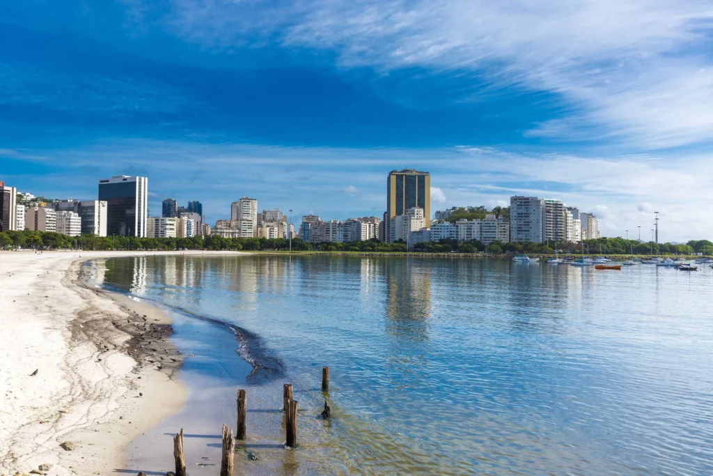 Imagem da praia de Botafogo, no Rio de Janeiro, mostra uma praia tranquila com ondas suaves, postes de madeira desgastados na areia e um horizonte urbano sob um céu azul brilhante. Árvores margeiam a orla ao fundo, contribuindo para o charme sereno do litoral.