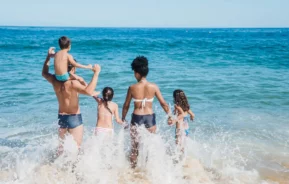 Imagem de uma família de cinco pessoas com trajes de banho entrando no mar em um dia ensolarado na praia.