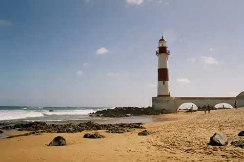 Imagem do Farol de Itapuã, em Salvador, em uma praia de areia perto do oceano, com pedras espalhadas ao longo da costa sob um céu azul claro.