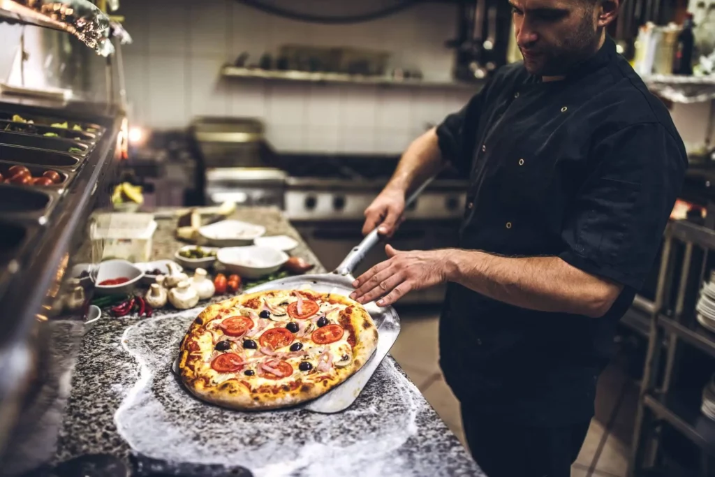 Imagem de UM chef com uniforme preto usando um cortador de pizza para fatiar uma pizza recém-assada em um balcão enfarinhado em uma cozinha profissional. Vários ingredientes e utensílios são visíveis ao fundo.