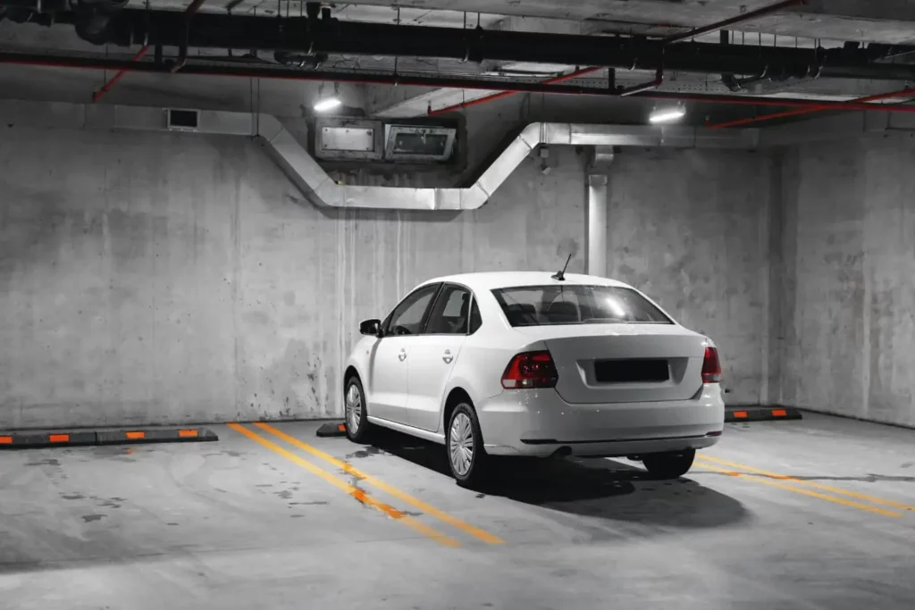 Imagem de um sedan branco estacionado em um estacionamento subterrâneo vazio com paredes e teto de concreto, vagas marcadas e tubos de ventilação visíveis.