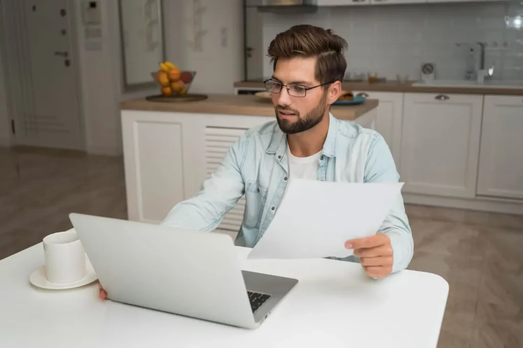 Imagem de um homem de óculos sentado na mesa de uma cozinha, olhando para a tela de um laptop enquanto segura uma folha de papel. Uma xícara e um pires estão sobre a mesa ao lado dele, e uma fruta pode ser vista ao fundo.