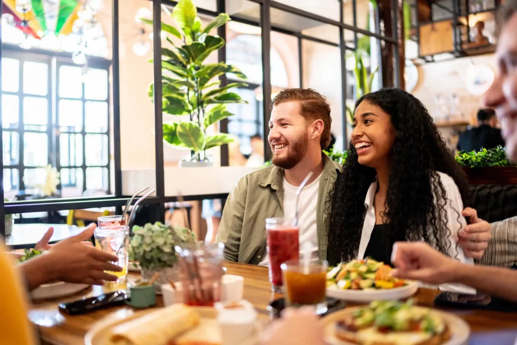 Imagem de um grupo de amigos sentados à mesa de um restaurante, sorrindo e saboreando comidas e bebidas. Duas pessoas no centro estão rindo, cercadas por pratos de salada e sanduíches. A atmosfera é animada.