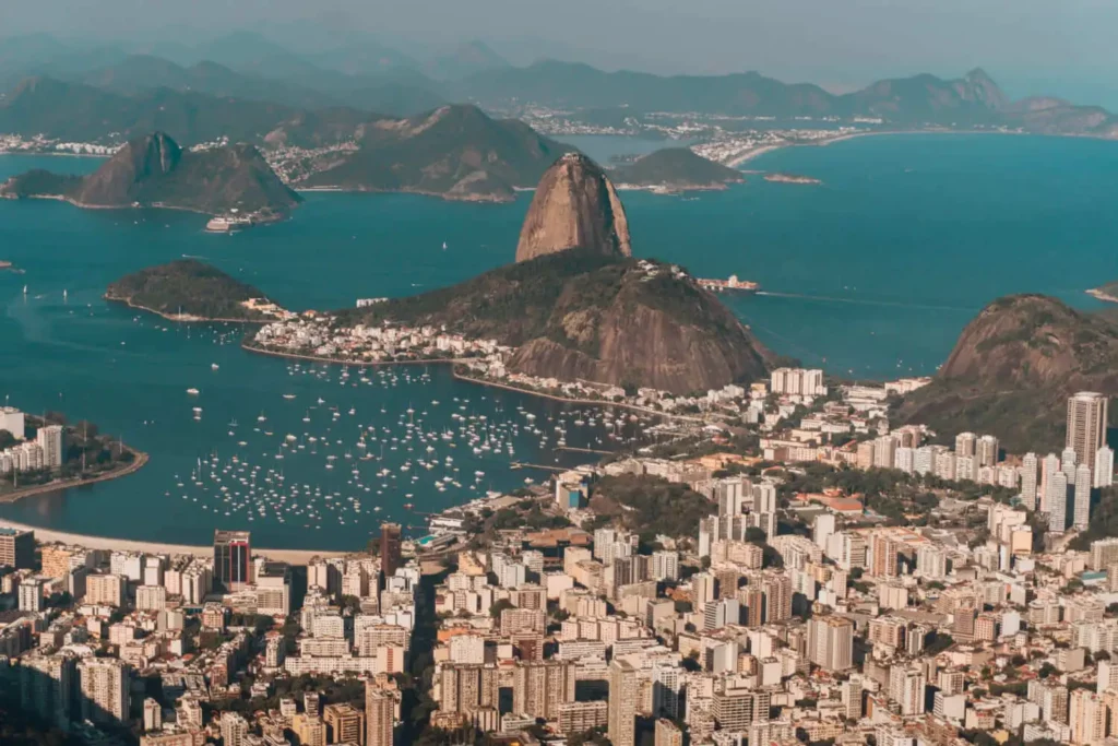 Imagem da vista aérea do Rio de Janeiro mostra o Pão de Açúcar, barcos na baía e prédios da cidade com montanhas distantes ao fundo.