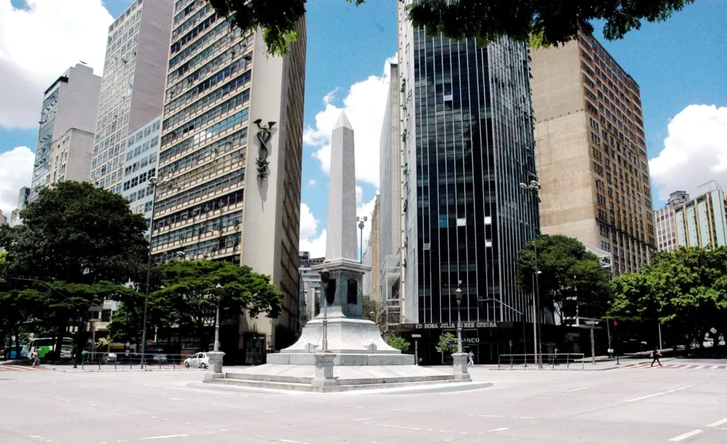 Imagem do Obelisco na Praça Sete de Setembro, em Belo Horizonte - Minas Gerais, cercado por edifícios altos, modernos e antigos, com árvores ao longo das bordas e um céu azul brilhante com nuvens espalhadas acima.