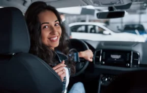 Imagem de uma mulher com longos cabelos escuros sentado no banco do motorista de um carro, sorrindo e olhando para trás por cima do ombro, com uma mão no volante. O interior do carro e alguns veículos do lado de fora são visíveis.