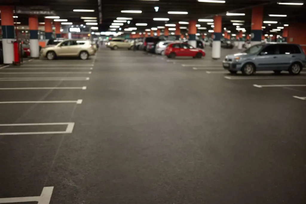 Imagem borrada de um estacionamento interno com vários carros estacionados em ambos os lados e muitas vagas vazias em primeiro plano. A garagem é bem iluminada com luzes no teto e pilares laranja.