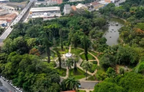 Vista aérea de um grande e exuberante parque urbano com muitas árvores, trilhas para caminhada, um pavilhão circular, um lago com barcos e ruas e edifícios da cidade ao redor.