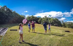 Um grupo de pessoas, incluindo crianças e adultos, empina pipas coloridas em um campo gramado cercado por árvores sob um céu azul brilhante com nuvens dispersas.